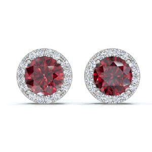 May include: A pair of stud earrings featuring a round, deep red gemstone surrounded by a halo of smaller, clear gemstones. The earrings have a silver-colored setting and are displayed against a white background.