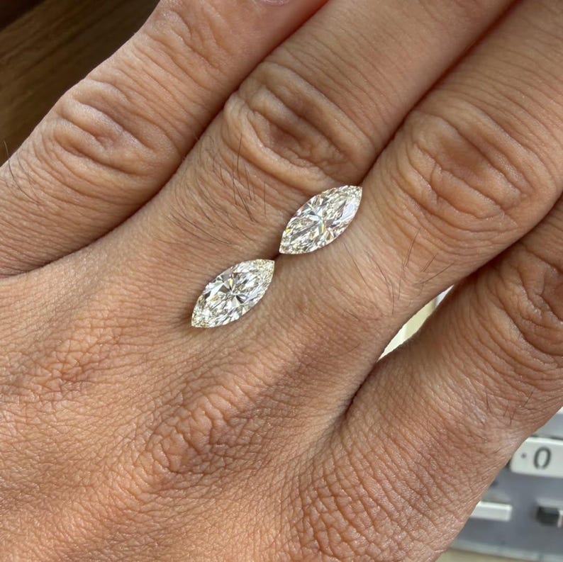 K&ouml;nnte beinhalten: Zwei Diamanten im Marquiseschliff, jeweils mit mehreren Facetten, werden gezeigt. Die Diamanten sind vor einem neutralen Hintergrund platziert, der ihre Brillanz hervorhebt. Die l&auml;ngliche Form der Diamanten ist ein wichtiges Merkmal, das Licht reflektiert und einen funkelnden Effekt erzeugt.
