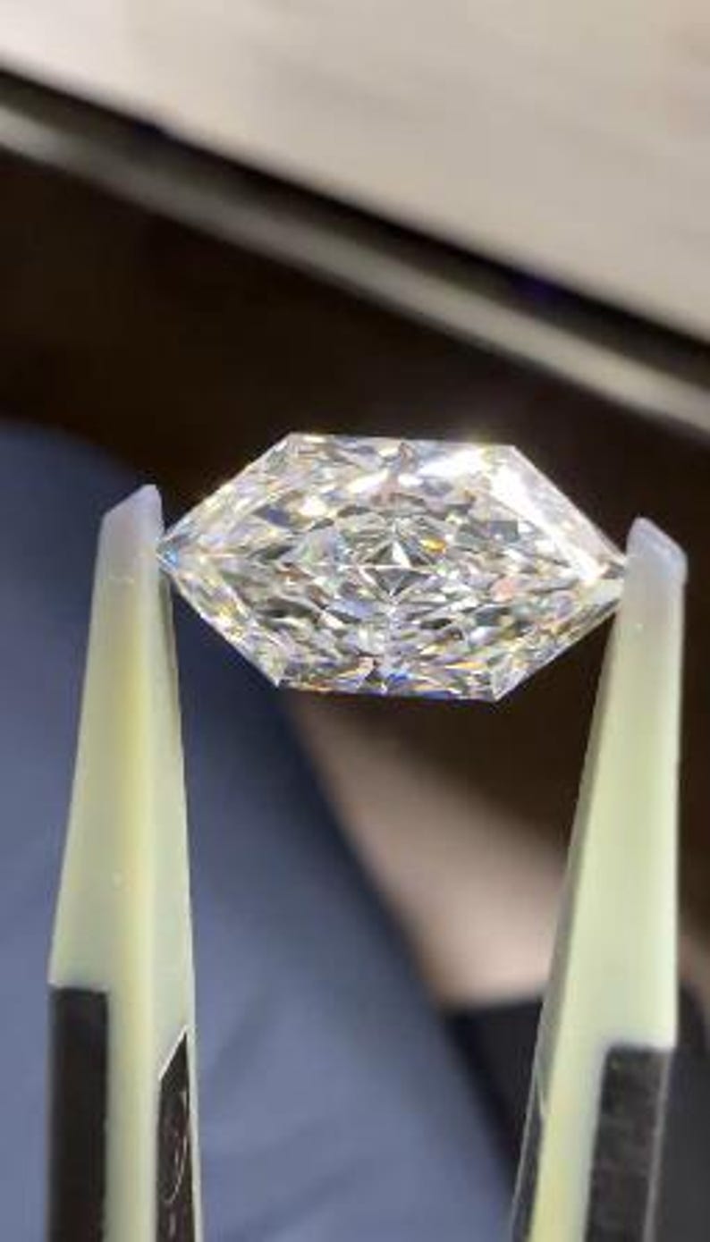 May include: A faceted, clear diamond held by tweezers. The diamond has a geometric shape with multiple facets that reflect light, creating a sparkling effect. The tweezers are light yellow and black.