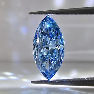 May include: A marquise-cut blue gemstone with a faceted surface that reflects light, creating bright highlights and deep azure tones. The stone is held vertically by dark metal tweezers, showcasing its elongated, pointed shape and clarity.