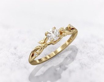 Könnte beinhalten: Ein goldener Ring mit einem zentralen, runden, klaren Edelstein. Der Ring hat ein zartes, von der Natur inspiriertes Design mit Blatt- und Weinranken-Details. Kleine, klare Akzentsteine sind entlang des Rings gesetzt. Der Ring ist mit "14K" markiert.