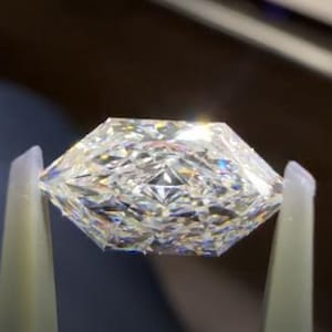 May include: A faceted, clear gemstone held by tweezers. The gemstone has a unique, geometric cut with multiple facets that reflect light, creating a sparkling effect. The shape is a modified octagon, with a flat top and a pointed bottom.