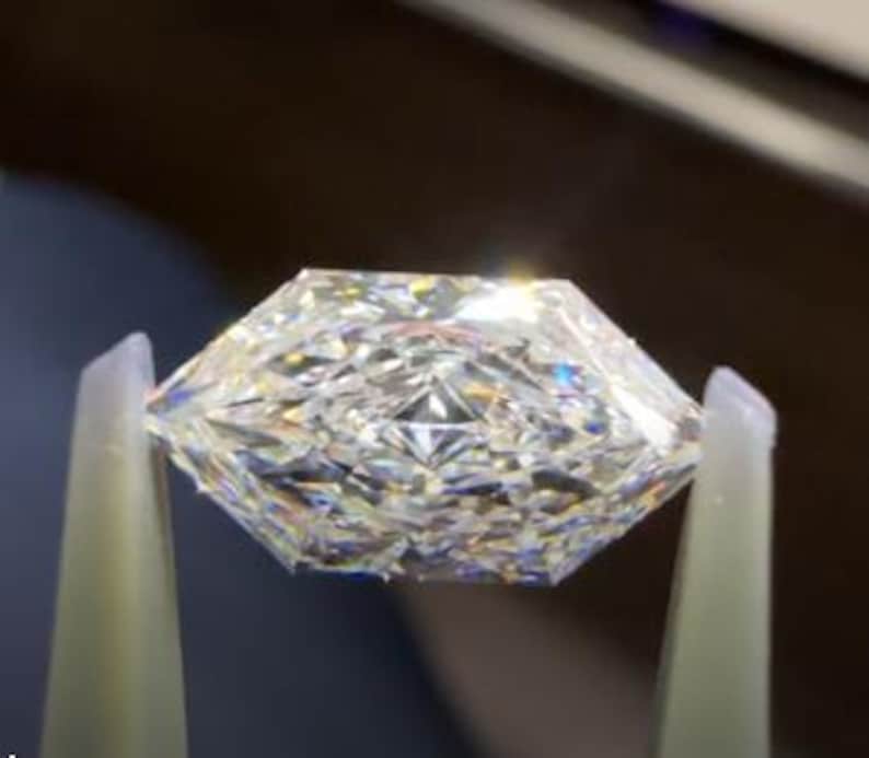 May include: A faceted, clear gemstone held by tweezers. The gemstone has a unique, geometric cut with multiple facets that reflect light, creating a sparkling effect. The shape is a modified octagon, with a flat top and a pointed bottom.