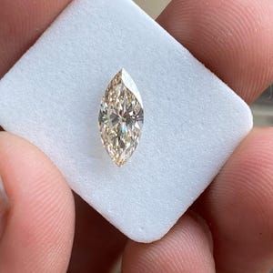 K&ouml;nnte beinhalten: Ein Diamant im Marquiseschliff mit einem warmen Champagnerton. Der Edelstein ist vor einem wei&szlig;en Hintergrund platziert, der seine l&auml;ngliche Form und seine brillanten Facetten hervorhebt. Der Diamant wird zwischen Fingern gehalten.