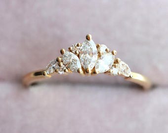 Puede incluir: Un anillo dorado con un grupo de piedras preciosas claras de talla marquesa y redonda. Las piedras están dispuestas en forma de corona. La banda del anillo es delgada y pulida.