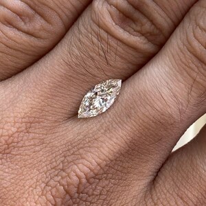 K&ouml;nnte beinhalten: Ein Diamant im Marquiseschliff mit einem hellen Champagnerton. Die l&auml;ngliche Form des Diamanten wird vor einem hautfarbenen Hintergrund gezeigt, der seine Facetten und seinen Glanz hervorhebt. Der Diamant wird in einer Hand gehalten.