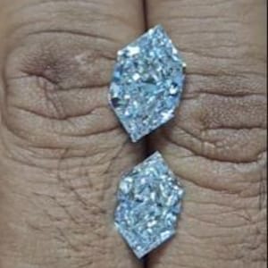 May include: Two clear, faceted gemstones with a unique, elongated shape. The gems are displayed against a neutral background, showcasing their brilliant sparkle and intricate cuts. The stones appear to be diamonds, reflecting light.