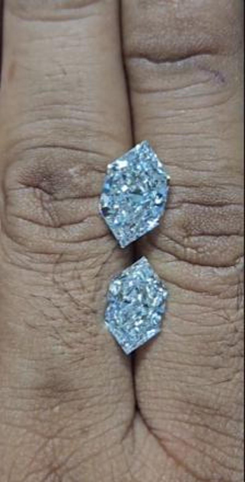 May include: Two clear, faceted gemstones with a unique, elongated shape. The gems are displayed against a neutral background, showcasing their brilliant sparkle and intricate cuts. The stones appear to be diamonds, reflecting light.