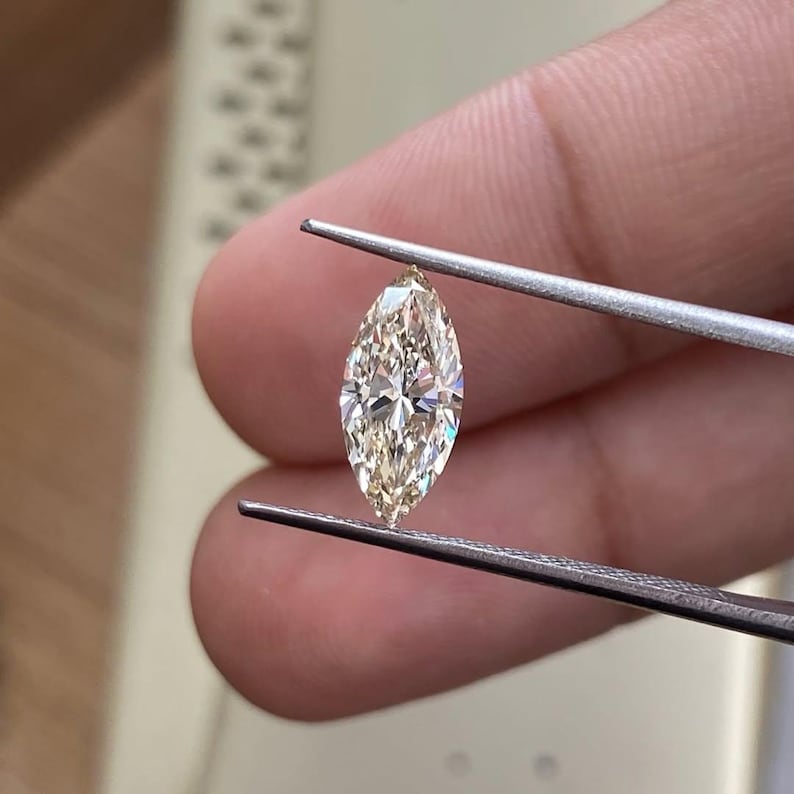 K&ouml;nnte beinhalten: Ein Diamant im Marquise-Schliff, der von einer Pinzette gehalten wird. Der Diamant hat eine hellchampagnerfarbene Farbe und einen Brillantschliff, der seine Facetten zur Geltung bringt. Die l&auml;ngliche Form wird zwischen den Metallspitzen gehalten.