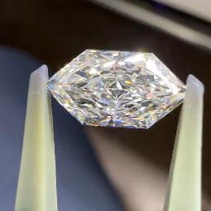 May include: A faceted, clear diamond held by tweezers. The diamond has a geometric shape with multiple facets that reflect light, creating a sparkling effect. The tweezers are light yellow and black.