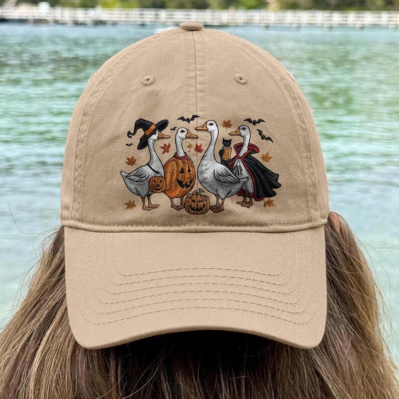 May include: Tan baseball cap featuring a Halloween-themed graphic. The design includes four geese dressed in costumes, including a witch hat, pumpkin, and vampire cape. Bats and fall leaves are also included in the design.