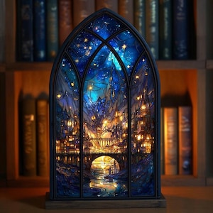 Può includere: Una luce notturna a forma di vetrata raffigurante una città fantasy sotto un cielo stellato. Il telaio della finestra è blu scuro e la città è illuminata da luci giallo caldo. La base è in legno.