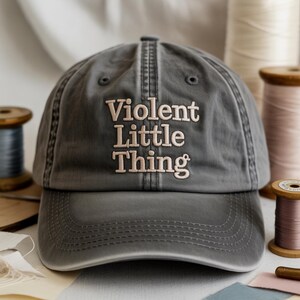 以下が含まれることがあります： 「Violent Little Thing」の文字が刺繍されたグレーの野球帽。つばはカーブしており、上部にステッチのデザインがあります。背景には糸巻きがあります。