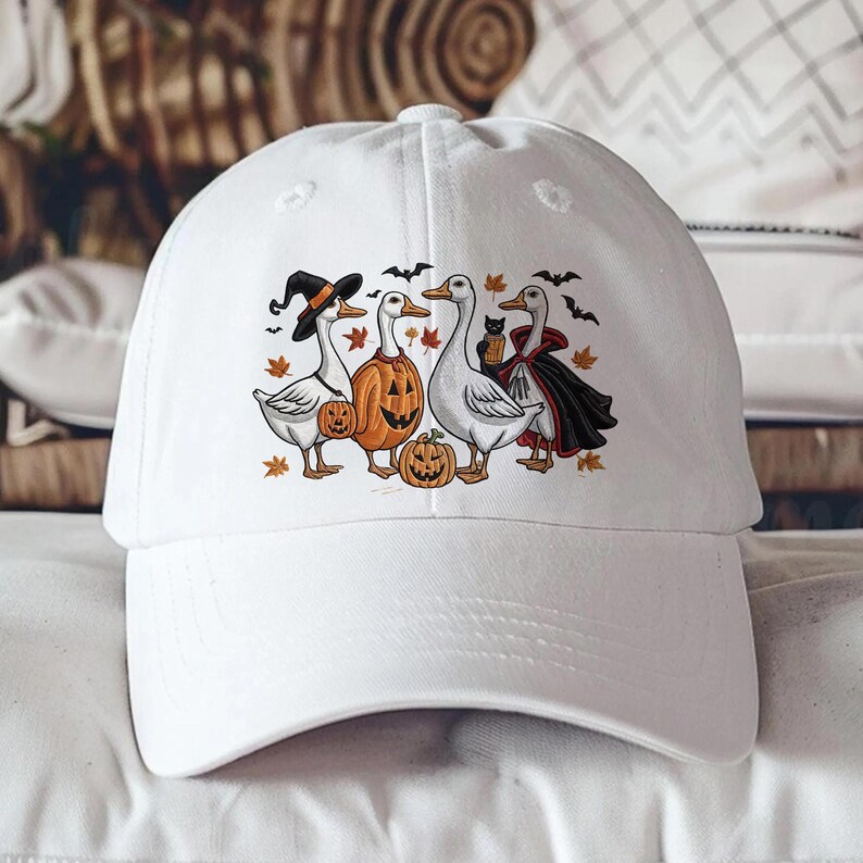 May include: White baseball cap featuring a Halloween-themed graphic. The design includes four geese dressed in costumes, pumpkins, bats, and autumn leaves. The geese are wearing a witch hat, a pumpkin head, a vampire cape, and holding a black cat.
