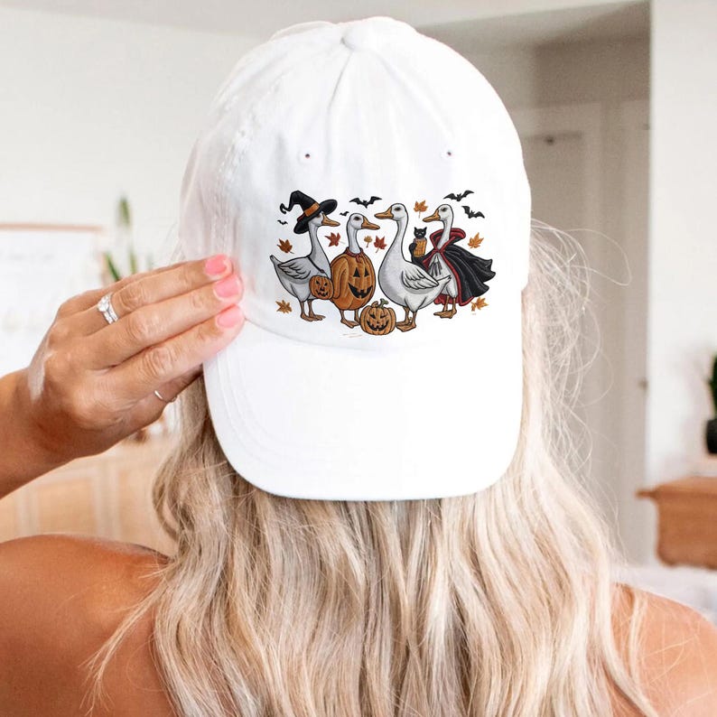May include: White baseball cap featuring a Halloween-themed graphic. The design includes four cartoon geese dressed in costumes, including a witch hat, pumpkin, and vampire cape. The geese are surrounded by bats and pumpkins.