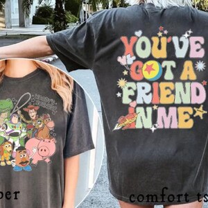 Puede incluir: Camiseta cómoda color pimienta con un gráfico de Toy Story en la parte delantera y la frase "You've Got a Friend in Me" en la parte trasera. El gráfico frontal incluye personajes como Buzz Lightyear y Woody.