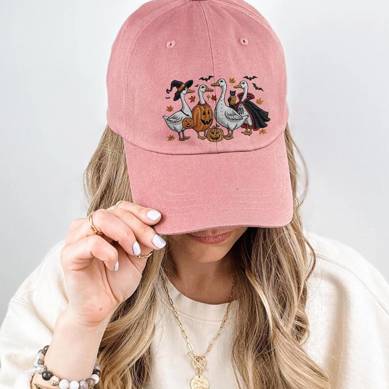 May include: A dusty rose baseball cap featuring a Halloween-themed graphic. The graphic depicts three geese dressed in Halloween costumes, including a witch, a pumpkin, and a vampire. The cap is made of a soft fabric.