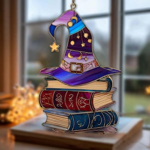 Op de afbeelding: Een gebrandschilderde heksenhoed met gouden accenten, manen en sterren, hangt bovenop een stapel van drie boeken. De hoed is paars, blauw en goud. De boeken zijn rood en blauw met gouden letters. Een gouden ster hangt aan de hoed.