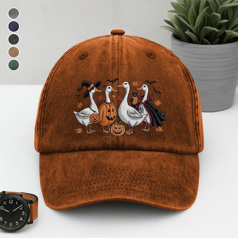 May include: A burnt orange baseball cap featuring an embroidered Halloween design. The design includes four geese dressed in costumes, pumpkins, and bats. The cap has a curved brim and a classic six-panel design.
