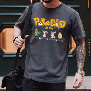 Puede incluir: Camiseta gris oscuro con el texto "PokeDad EST. 2026" en amarillo y naranja. Debajo, imágenes de dibujos animados de personajes de Pokémon y nombres. Estilo casual.