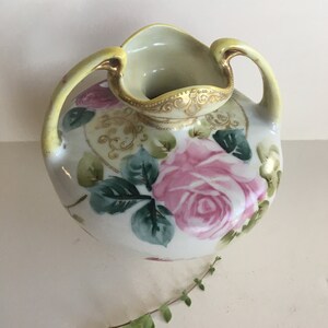 Könnte beinhalten: Eine weiße Porzellanvase mit zwei Henkeln, verziert mit rosa Rosen und grünen Blättern. Die Vase hat einen goldenen Rand am oberen Rand und eine hellgelbe Glasur.