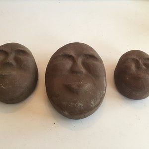 K&ouml;nnte beinhalten: Drei braune Keramikskulpturen von Gesichtern mit geschlossenen Augen. Die Skulpturen sind unterschiedlich gro&szlig; und in einer Reihe vom gr&ouml;&szlig;ten zum kleinsten angeordnet.