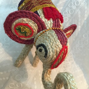 May include: A hand-crafted elephant figurine made of woven rope and decorated with colorful fabric and gold accents. The elephant has a red, white, and blue patterned head with a gold trim and a long, curled trunk.