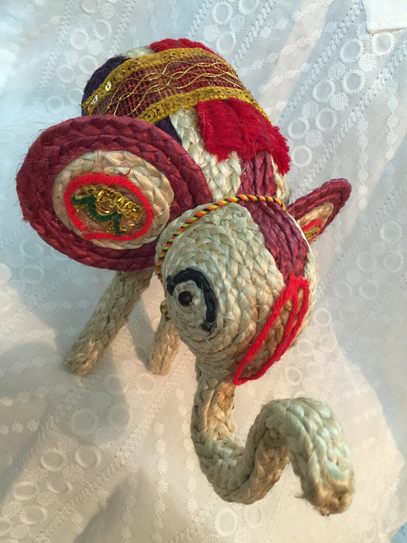 May include: A hand-crafted elephant figurine made of woven rope and decorated with colorful fabric and gold accents. The elephant has a red, white, and blue patterned head with a gold trim and a long, curled trunk.