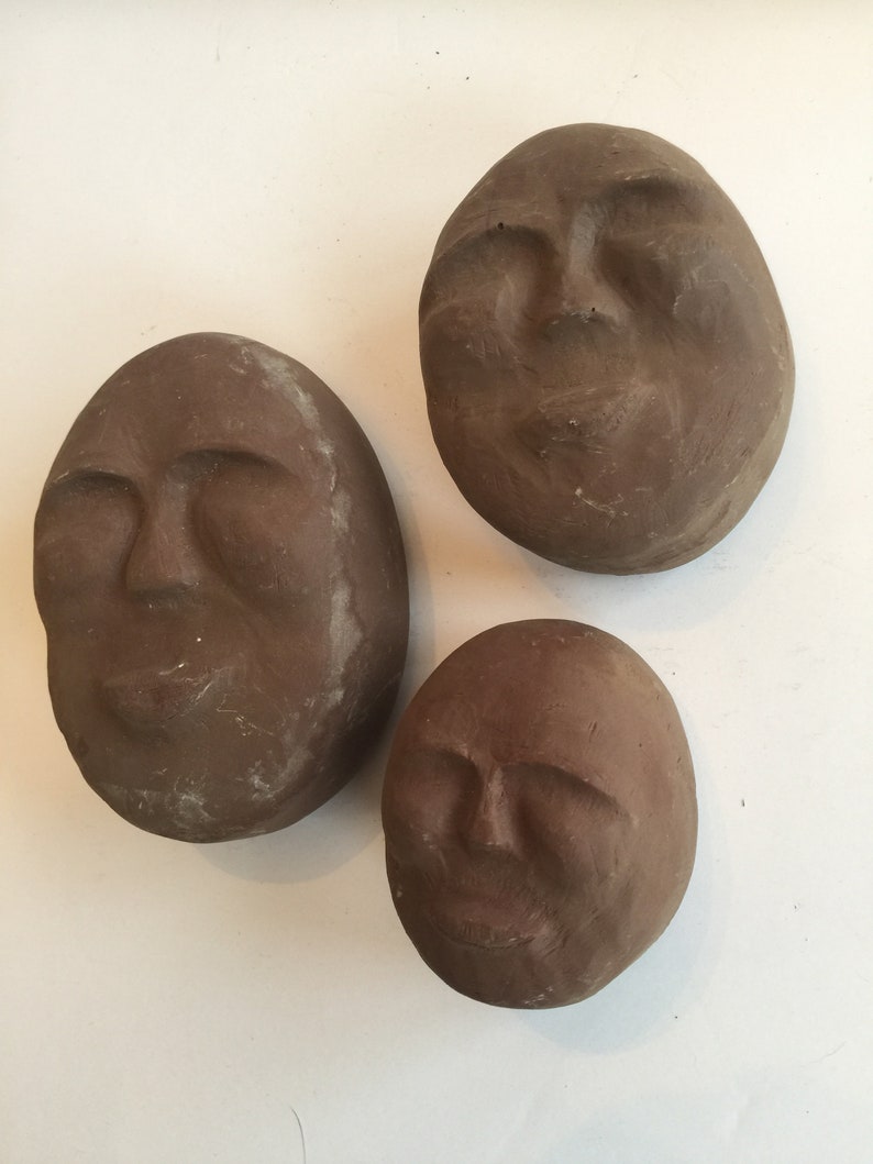 K&ouml;nnte beinhalten: Drei braune Tonskulpturen von Gesichtern mit geschlossenen Augen. Die Skulpturen sind rund und haben eine glatte, strukturierte Oberfl&auml;che.