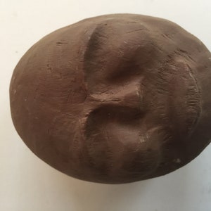 K&ouml;nnte beinhalten: Eine braune Tonskulptur eines Gesichts mit geschlossenem Mund und Augen. Die Skulptur ist rund und hat eine glatte Oberfl&auml;che.
