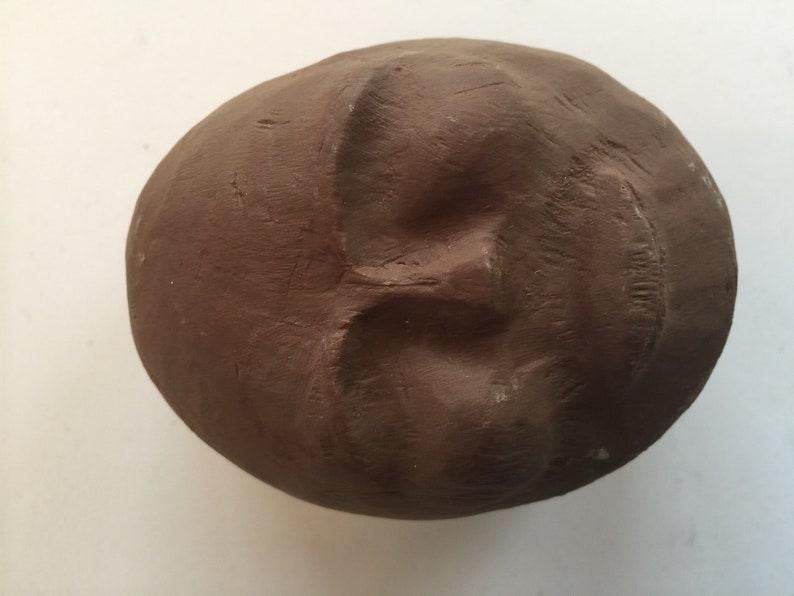 K&ouml;nnte beinhalten: Eine braune Tonskulptur eines Gesichts mit geschlossenem Mund und Augen. Die Skulptur ist rund und hat eine glatte Oberfl&auml;che.