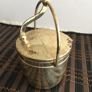 May include: A silver-plated, round, lidded container with a handle. The container is sitting on a brown woven mat.