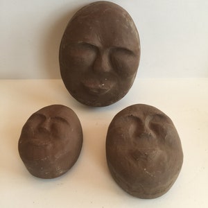 K&ouml;nnte beinhalten: Drei braune Keramikskulpturen von Gesichtern mit geschlossenen Augen. Die Skulpturen sind oval geformt und haben eine strukturierte Oberfl&auml;che.