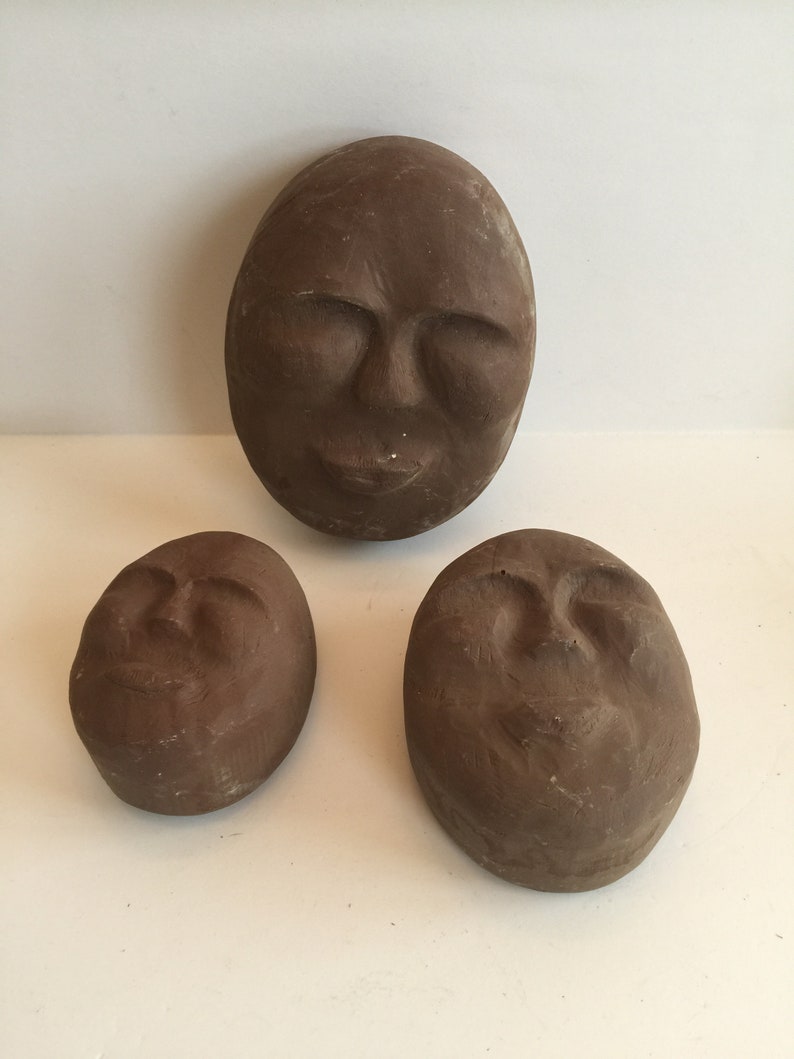 K&ouml;nnte beinhalten: Drei braune Keramikskulpturen von Gesichtern mit geschlossenen Augen. Die Skulpturen sind oval geformt und haben eine strukturierte Oberfl&auml;che.