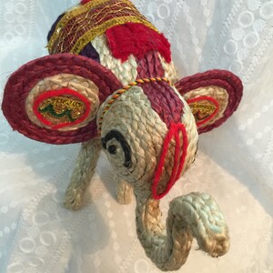 May include: A hand-crafted elephant figurine made of woven jute and decorated with colorful fabric and gold thread. The elephant has large, round ears and a long trunk.