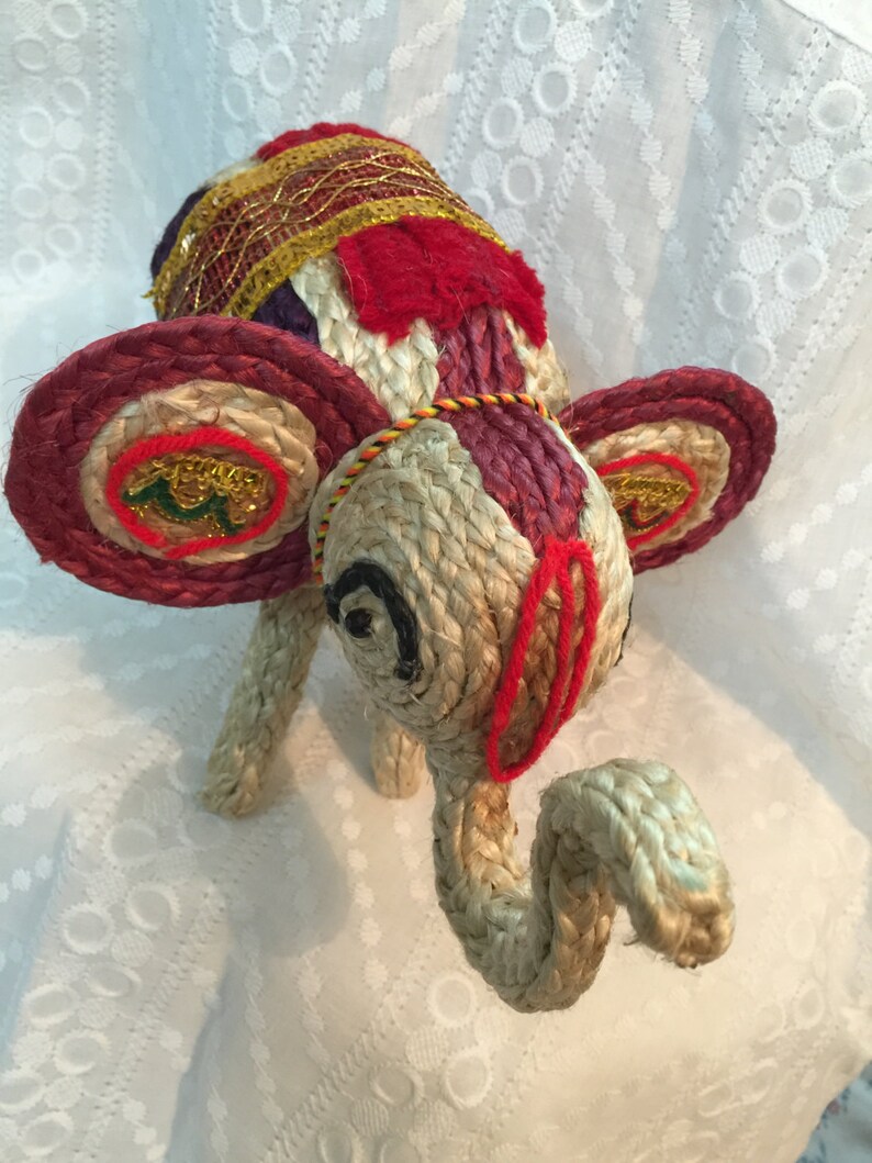 May include: A hand-crafted elephant figurine made of woven jute and decorated with colorful fabric and gold thread. The elephant has large, round ears and a long trunk.