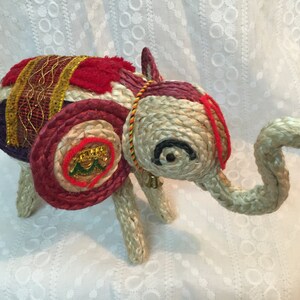 May include: A hand-crafted elephant figurine made of woven straw and decorated with colorful fabric and sequins. The elephant has a red, white, and gold saddle and a long, curled trunk.
