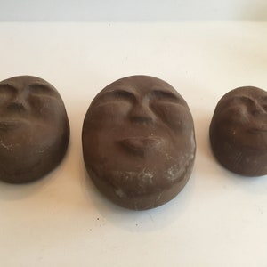 K&ouml;nnte beinhalten: Drei braune Keramikskulpturen von Gesichtern mit geschlossenen Augen und leicht ge&ouml;ffnetem Mund. Die Skulpturen sind unterschiedlich gro&szlig; und in einer Reihe vom gr&ouml;&szlig;ten zum kleinsten angeordnet.