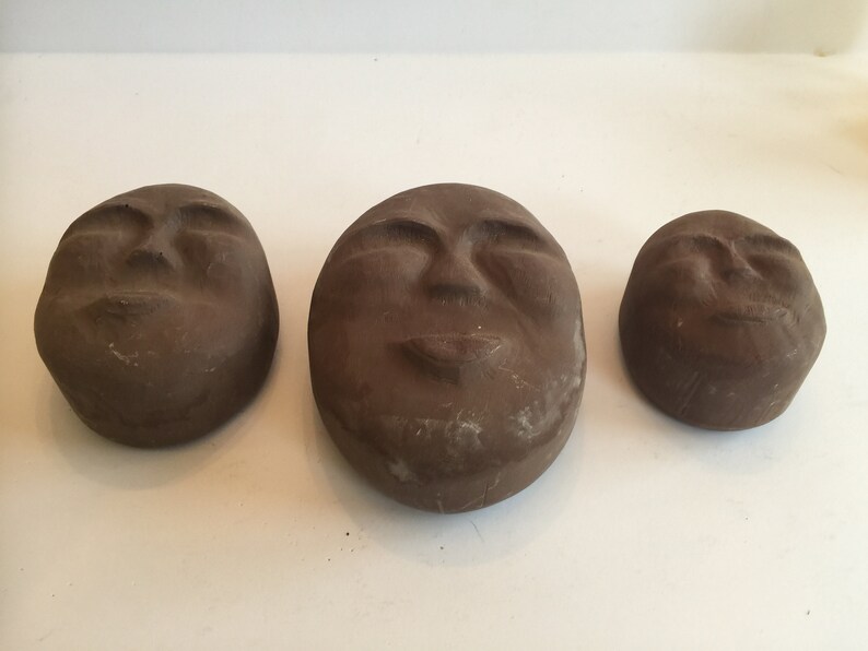 K&ouml;nnte beinhalten: Drei braune Keramikskulpturen von Gesichtern mit geschlossenen Augen und leicht ge&ouml;ffnetem Mund. Die Skulpturen sind unterschiedlich gro&szlig; und in einer Reihe vom gr&ouml;&szlig;ten zum kleinsten angeordnet.