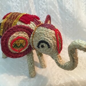 May include: A hand-crafted elephant figurine made of woven straw, decorated with colorful fabric and gold accents. The elephant has a red, white, and gold saddle and a bell hanging from its neck.
