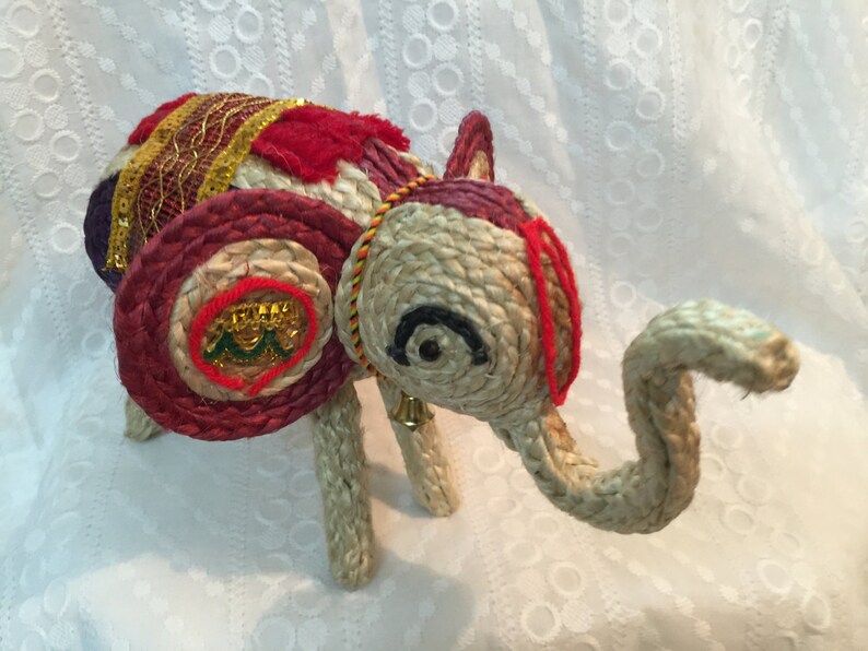 May include: A hand-crafted elephant figurine made of woven straw, decorated with colorful fabric and gold accents. The elephant has a red, white, and gold saddle and a bell hanging from its neck.