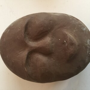 K&ouml;nnte beinhalten: Eine braune Tonskulptur eines Gesichts mit geschlossenen Augen und leicht ge&ouml;ffnetem Mund.