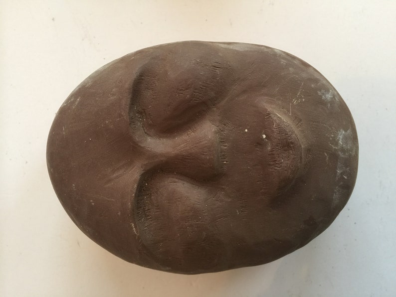 K&ouml;nnte beinhalten: Eine braune Tonskulptur eines Gesichts mit geschlossenen Augen und leicht ge&ouml;ffnetem Mund.