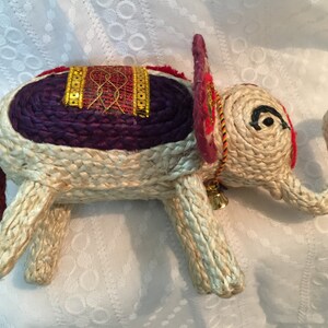 May include: A woven straw elephant figurine with a purple and red saddle, gold trim, and a small bell. The elephant has a curled trunk and black eyes.