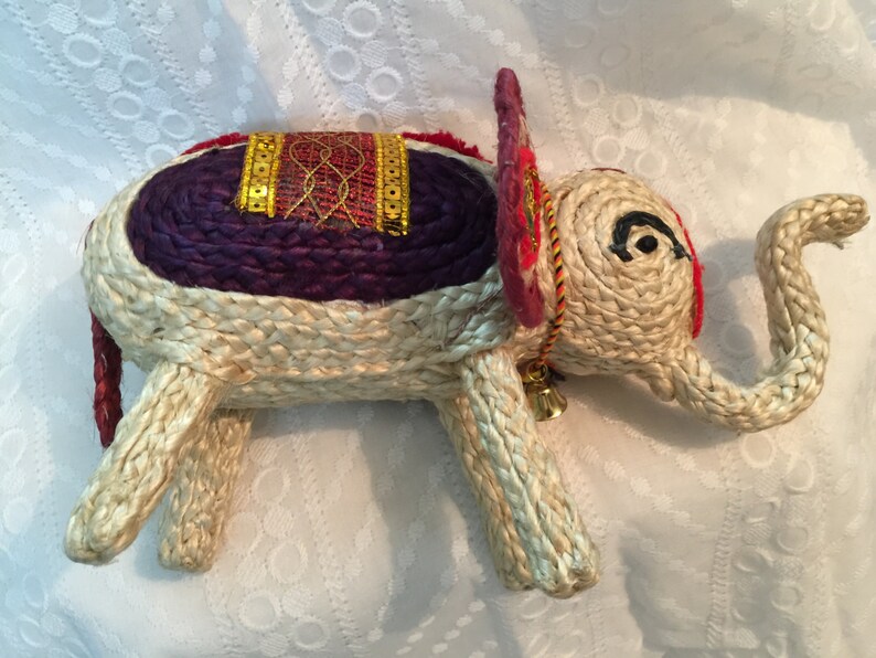 May include: A woven straw elephant figurine with a purple and red saddle, gold trim, and a small bell. The elephant has a curled trunk and black eyes.