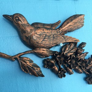 May include: A copper-colored bird sculpture perched on a branch with delicate flowers. The bird has a detailed, textured design and is a decorative wall hanging.
