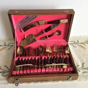 May include: A wooden box with a pink velvet interior containing a set of gold-tone silverware with brown handles. The box has a label that reads "Tony's Gems".