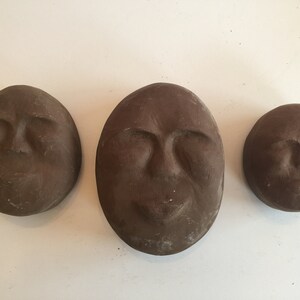K&ouml;nnte beinhalten: Drei braune Tonskulpturen von Gesichtern mit geschlossenen Augen. Die Skulpturen sind unterschiedlich gro&szlig; und in einer Reihe angeordnet.