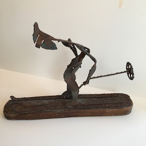 Könnte beinhalten: Eine rostige Metallsculptur eines Skifahrers auf einem Holzsockel. Der Skifahrer ist in der Luft dargestellt, mit einem Ski nach vorne gestreckt und dem anderen Ski dahinter. Die Arme des Skifahrers sind ausgestreckt, und die Skulptur besteht aus verschiedenen Metallteilen, die miteinander verschweißt wurden.