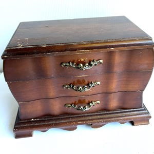 May include: A dark brown wooden jewelry box featuring a rectangular top, three horizontal drawers with wavy fronts, and ornate brass-toned metal handles. The piece stands on a decorative base with feet and shows visible wear on the wood finish.